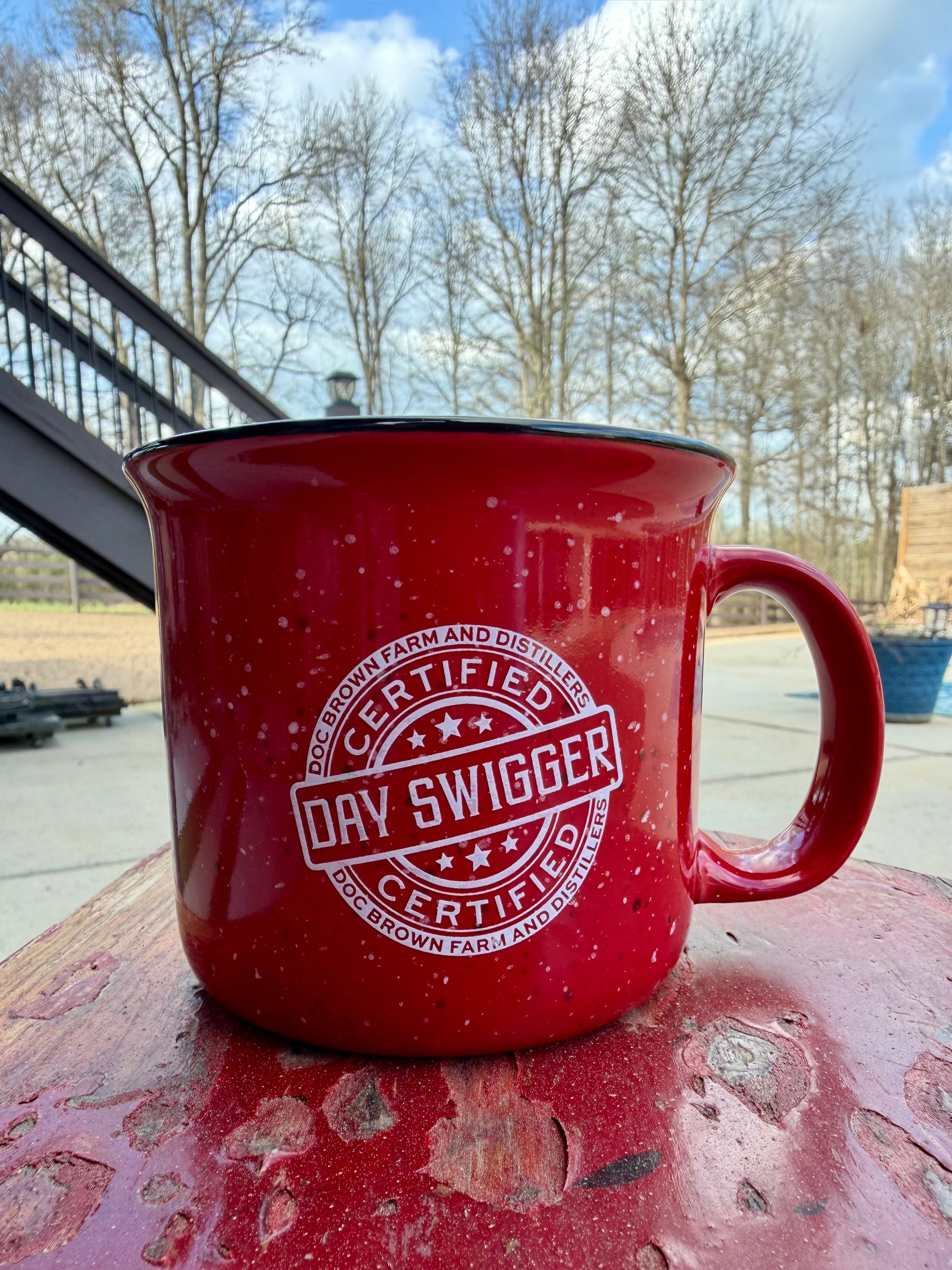 Red mug with 'Certified Day Swigger' logo on a wooden surface outdoors.